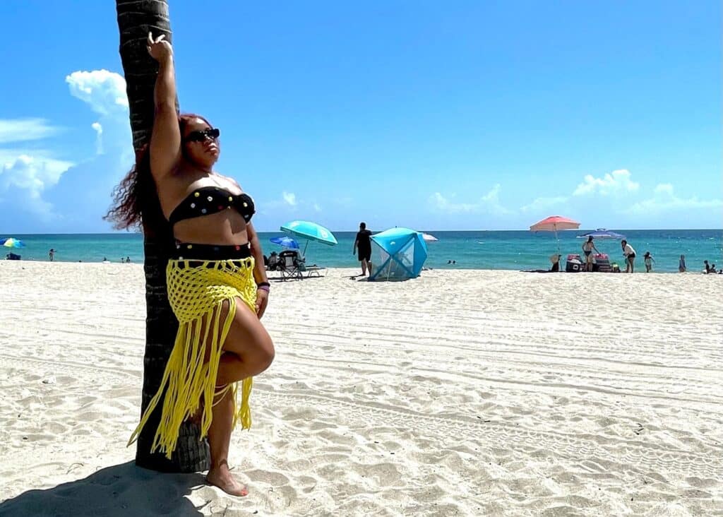 Me in a black bikini and yellow crochet coverup on the beach.