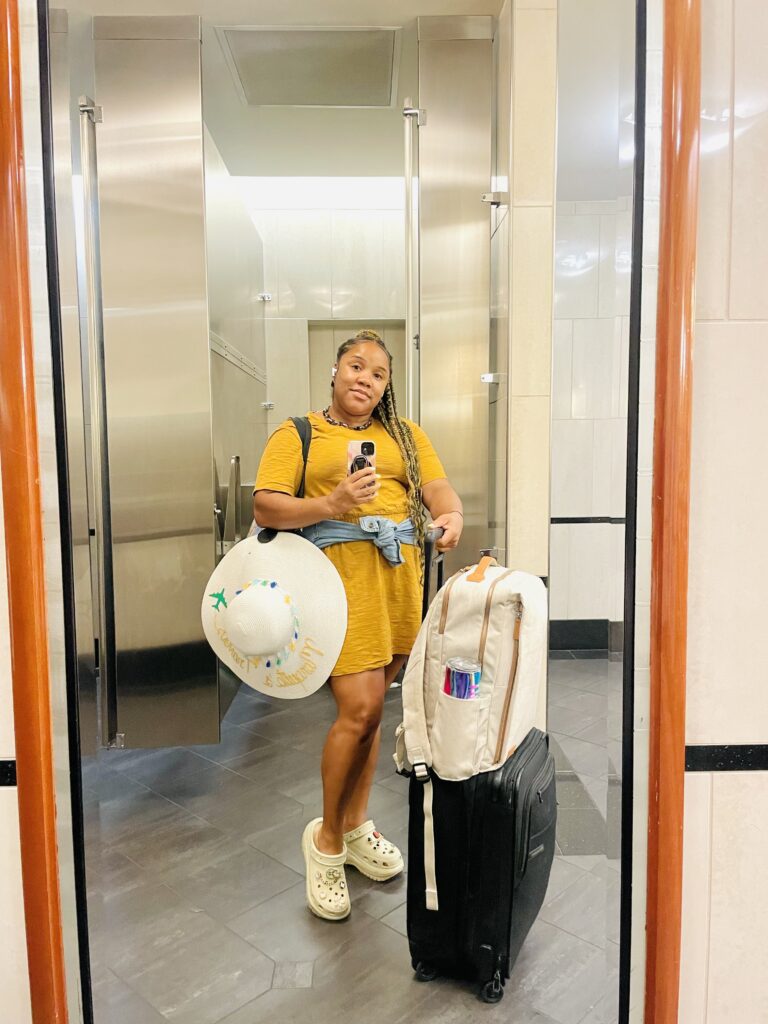 Airport bathroom selfie. I'm dressed in a sunflower yellow, above the knee dress, with a sweatshirt tied around my waist. Next to me is my carry-on luggae and travel backpack- as well as my oversize beach hat. 